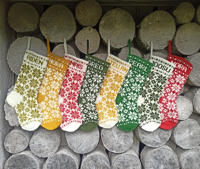 May include: Eight knitted Christmas stockings featuring a repeating snowflake pattern in various colors including olive green, mustard yellow, red, and white. Each stocking is personalized with a name at the top: Aiden, Nora, Nais, Greg, Stellan, Pearl, Joosu, and Nora.