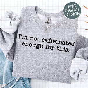 May include: A grey sweatshirt with the phrase "I'm not caffeinated enough for this." A silver necklace with a sun pendant is on the shirt. The sweatshirt is folded on top of blue denim.