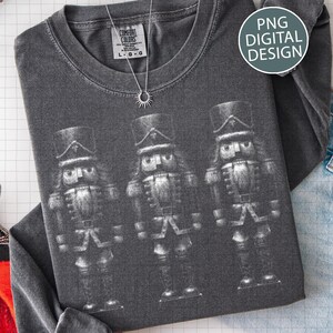 May include: Dark gray long-sleeve shirt featuring a graphic of three nutcrackers. The shirt has a crew neck and a tag that says "Comfort Colors". A silver necklace with a sunburst pendant is also visible. The words "PNG DIGITAL DESIGN" are in a circle.