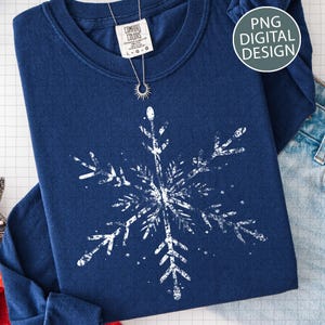May include: A navy blue long-sleeve sweatshirt featuring a large white snowflake design. The shirt has a crew neck and a tag that reads "Comfort Colors". A silver necklace with a sun pendant is draped over the shirt. The words "PNG DIGITAL DESIGN" are in a circle.