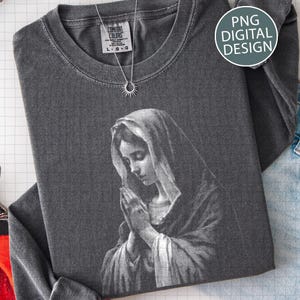 May include: A charcoal gray long-sleeve shirt featuring a black and white image of a praying woman. The shirt has a crew neck and a tag that reads "Comfort Colors". A silver necklace with a sun pendant is also visible. The image also includes the text "PNG DIGITAL DESIGN".