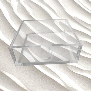 May include: A clear, square acrylic holder with a U-shaped cutout on the front panel. The container features four vertical sides and an open top, constructed from transparent material with visible seams at the corners and a smooth, polished finish.