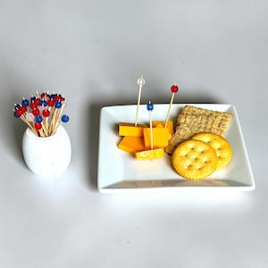 Op de afbeelding: Een witte keramische houder met houten tandenstokers met kleine rode, witte en blauwe kralen. Daarnaast staat een vierkant wit bordje met blokjes oranje cheddar, rechthoekige tarwecrackers en ronde goudkleurige crackers.