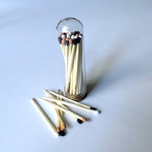 Puede incluir: Un recipiente cilíndrico de vidrio transparente con base de corcho lleno de cerillas de madera con puntas negras, blancas y color canela. Varias cerillas sueltas con puntas de colores similares están esparcidas frente al recipiente.