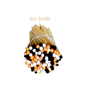 May include: A bundle of wooden matches tied together with a clear string, featuring match heads dipped in a mix of black, orange, and white colors. The text "boo bundle" is printed in a light brown font above the matchsticks.