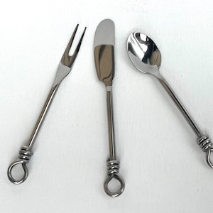 May include: Set of three stainless steel serving utensils: a two-tine fork, a butter knife, and a spoon. Each utensil has a long, thin handle with a coiled wire design near the end and a loop at the very end.