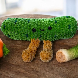 May include: A handmade green crochet pickle with black button eyes and orange feet sits on a wooden board. Fresh vegetables, including lettuce, tomatoes, and a green and white vegetable, are also on the board.