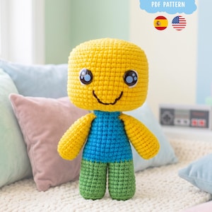 Pode incluir: Uma boneca de crochê com uma grande cabeça quadrada amarela, um tronco azul e pernas verdes. A boneca tem olhos bordados pretos com destaques brancos e um sorriso simples costurado em preto. Uma nuvem azul com o texto PDF PATTERN é visível.
