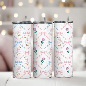 Op de afbeelding: Drie witte roestvrijstalen bekers met een patroon van roze en blauwe strikken, witte ganzen en paarse bloemen.