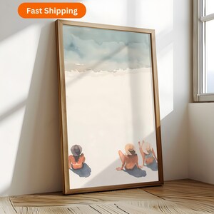 May include: A framed watercolour print depicting a beach scene. Three figures in swimwear and hats are sunbathing on the sand. The artwork features a light blue sky and ocean. The frame is made of wood.