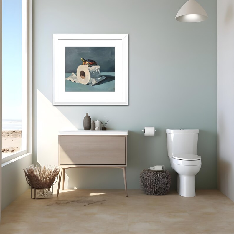 May include: A framed artwork depicts a turtle atop a roll of toilet paper. The artwork is in a white frame and hangs above a bathroom vanity. The bathroom has a toilet, a small stool, and a window with a view of the beach.