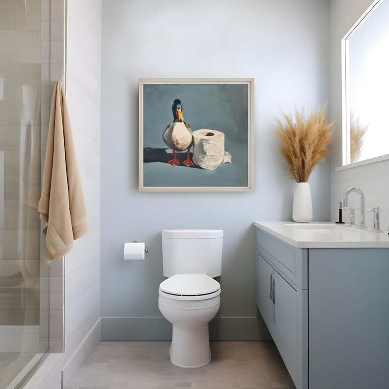 May include: A framed painting of a duck and a roll of toilet paper hangs above a white toilet in a bathroom. The painting has a light blue background. The bathroom has a light blue vanity and a shower with a beige towel.