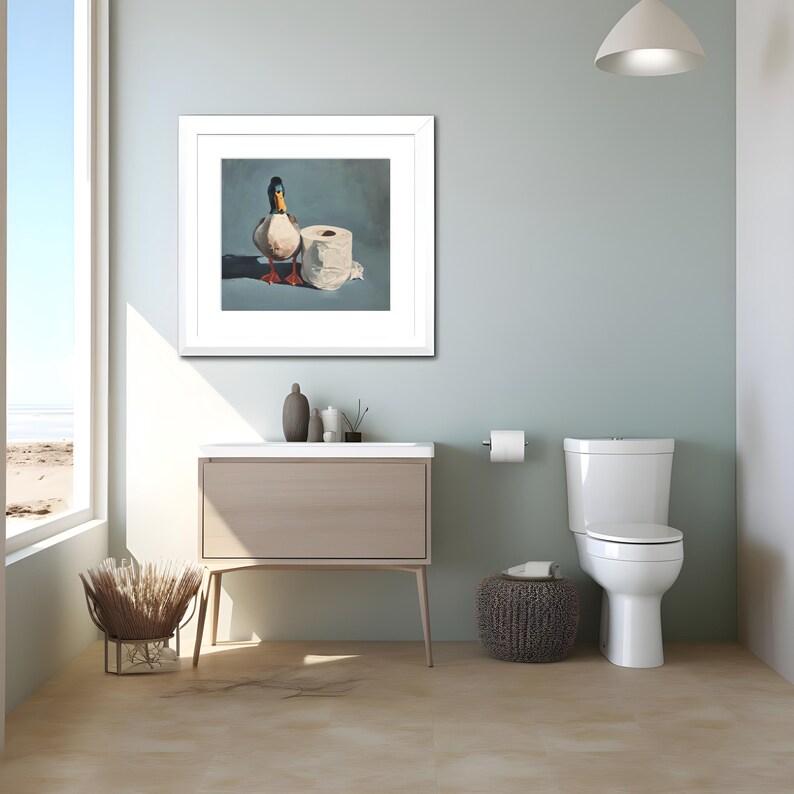 May include: A framed artwork depicting a duck and a roll of toilet paper. The artwork is displayed in a modern bathroom setting with a white toilet, a light wood vanity, and a woven stool. The walls are painted a light blue.