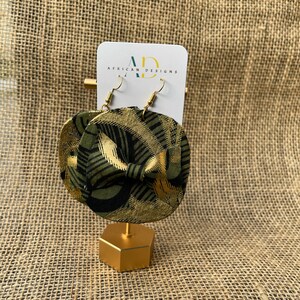 May include: A pair of gold and green fabric earrings with a geometric pattern. The earrings are on a gold stand and have a white card behind them that says "African Designs".