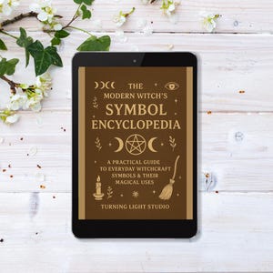 Könnte beinhalten: Ein Tablet zeigt "The Modern Witch's Symbol Encyclopedia" mit braunem Einband und goldener Schrift. Das Cover zeigt Hexensymbole wie Halbmond, Pentagramm und Besen. Das Tablet liegt auf einer weißen Holzoberfläche mit floralen Akzenten.