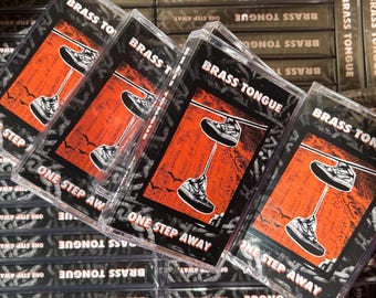 May include: A stack of cassette tapes with the band name "Brass Tongue" and the album title "One Step Away" printed on the front. The tapes have a black background with orange and white graphics.
