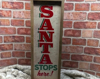 May include: A tall rectangular wooden sign with a light cream background and a dark wood frame. The sign features the red text SANTA in a bold font, followed by the green text STOPS and the red cursive text here! with an exclamation point.