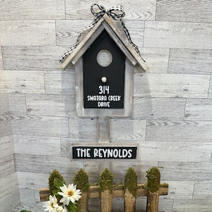 May include: A wooden birdhouse with a black chalkboard sign that reads "314 Swatara Creek Drive" and a smaller sign that reads "The Reynolds". The birdhouse is decorated with a black and white checkered ribbon. The birdhouse is sitting on a wooden fence with moss and white flowers. Two white ceramic birds are sitting on a black metal stand in front of the fence.