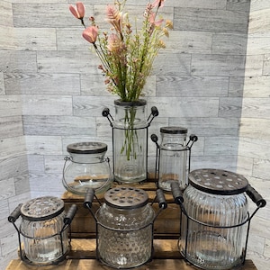 Peut inclure: Six vases en verre transparent avec des cadres et des couvercles en métal. Les vases ont une variété de textures, y compris côtelées, texturées et lisses. Les vases sont disposés sur une surface en bois. Un bouquet de fleurs roses est dans l'un des vases.