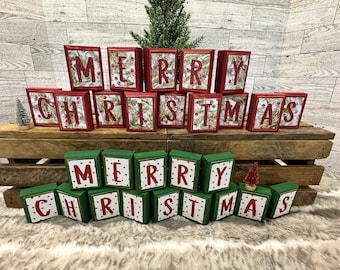 May include: Wooden blocks spelling "MERRY CHRISTMAS" in red letters. The blocks are red and green, with a festive design. The letters are set against a background of greenery and berries, perfect for holiday decor.