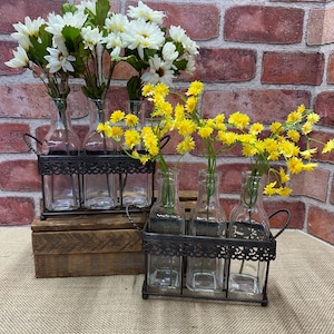 Peut inclure: Deux ensembles de bouteilles en verre transparent dans des cages en fil métallique avec des fleurs blanches et jaunes. Les cages ont un design décoratif et sont posées sur une surface en bois.