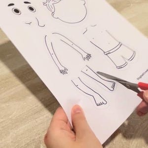 Puede incluir: Una mu&ntilde;eca de papel imprimible recortada de una persona con la cara en blanco, brazos, piernas y torso. La persona lleva pantalones cortos. La mu&ntilde;eca de papel se est&aacute; recortando con unas tijeras.