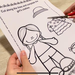 Puede incluir: Una hoja de actividad imprimible en blanco y negro con el contorno de una ni&ntilde;a y piezas de ropa para recortar. El texto en la hoja dice "Corta a lo largo de las l&iacute;neas y completa la ropa de la ni&ntilde;a."