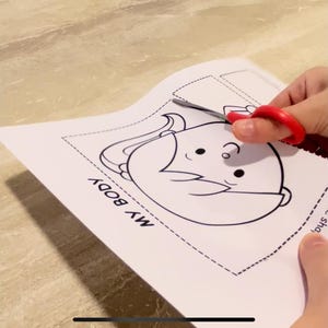 Puede incluir: Una actividad de manualidades imprimible en blanco y negro para ni&ntilde;os. La imprimible presenta un personaje de dibujos animados con el texto "MY BODY" en la p&aacute;gina. Un par de tijeras rojas est&aacute; recortando el personaje.