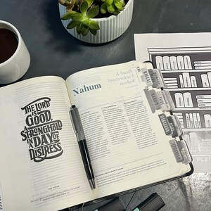 May include: An open Bible with a black pen and a cup of coffee. The Bible is open to the book of Nahum. The page has the text "The Lord is good, a stronghold in a day of distress". There are several grey tabs on the side of the Bible with the names of different books of the Bible. A sheet of paper with a drawing of a bookshelf with the names of different books of the Bible is on the table.