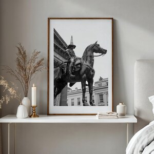 Op de afbeelding: Zwart-wit ingelijste print van een standbeeld van een persoon te paard. Het kunstwerk wordt tentoongesteld op een witte tafel met decoratieve items, waaronder een kaars en vazen. De kamer heeft neutrale tinten en een bed.