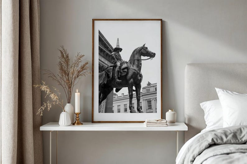 Op de afbeelding: Zwart-wit ingelijste print van een standbeeld van een persoon te paard. Het kunstwerk wordt tentoongesteld op een witte tafel met decoratieve items, waaronder een kaars en vazen. De kamer heeft neutrale tinten en een bed.