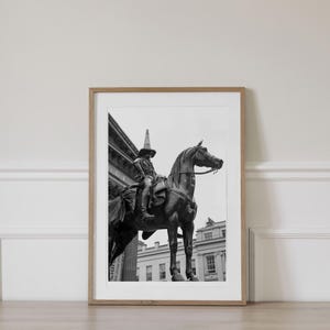 Op de afbeelding: Ingelijste zwart-wit print van een standbeeld van een persoon te paard. Het standbeeld staat voor een gebouw. De persoon draagt een hoed. De print zit in een houten lijst.