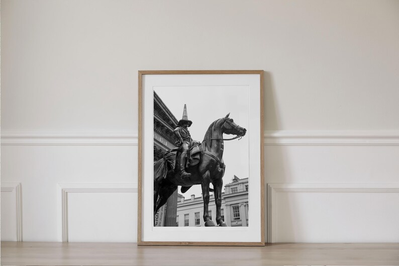 Op de afbeelding: Ingelijste zwart-wit print van een standbeeld van een persoon te paard. Het standbeeld staat voor een gebouw. De persoon draagt een hoed. De print zit in een houten lijst.