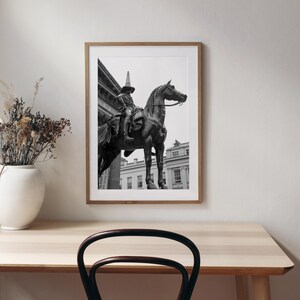 Op de afbeelding: Ingelijste zwart-wit print van een standbeeld van een persoon te paard. Het standbeeld staat voor een gebouw met zuilen. De print zit in een houten lijst en wordt tentoongesteld op een lichtgekleurde houten tafel. Een witte vaas met droogbloemen staat op de tafel.