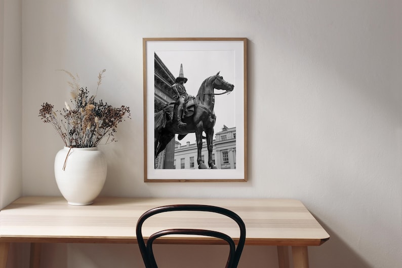 Op de afbeelding: Ingelijste zwart-wit print van een standbeeld van een persoon te paard. Het standbeeld staat voor een gebouw met zuilen. De print zit in een houten lijst en wordt tentoongesteld op een lichtgekleurde houten tafel. Een witte vaas met droogbloemen staat op de tafel.