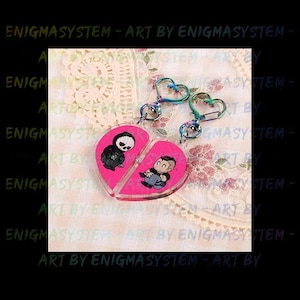 May include: Two pink heart-shaped keychains with a black and white illustration of a skeleton and a person on each side. The keychains are attached to rainbow-coloured metal split rings.