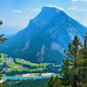 May include: A panoramic view of a mountain range with a river and a golf course in the foreground. The mountain is a light blue color with a white peak. The river is a light blue color and the golf course is a green color.