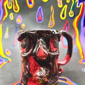 Pode incluir: Caneca de cerâmica artesanal com design semelhante a um rosto. A caneca apresenta um esmalte vermelho e preto com uma superfície texturizada. A alça é vermelha e o fundo tem padrões de tinta coloridos e gotejantes. Uma peça única.