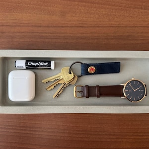 May include: A rectangular, light gray tray holds everyday items. Included are a white AirPods case, a tube of ChapStick, a set of keys, a navy blue key fob with a red button, and a wristwatch with a brown leather band.