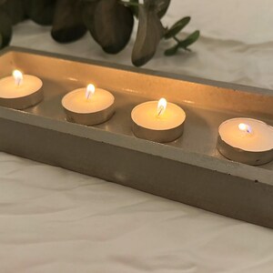Może przedstawiać: Prostokątna betonowa taca mieści cztery zapalone świece typu tealight. Świece mają miękki, ciepły blask. Taca jest w neutralnym szarym kolorze, a tło to biała powierzchnia z zielenią.