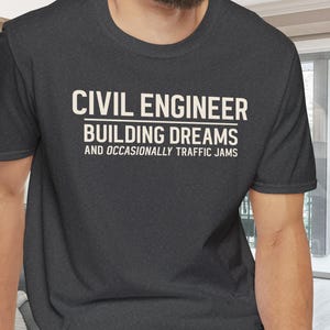 May include: Dark gray t-shirt with the text "CIVIL ENGINEER BUILDING DREAMS AND OCCASIONALLY TRAFFIC JAMS" in white block letters. The shirt is made of a soft material and has short sleeves and a crew neck.
