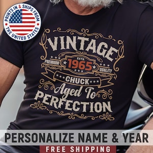 May include: A black t-shirt with a vintage-style graphic design. The design features the text "Vintage" with the year "1965" and the name "Chuck". The text "Aged to Perfection" is also included in the design.