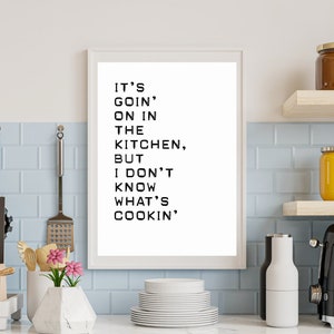 May include: A white framed print with black text that reads "IT'S GOIN' ON IN THE KITCHEN, BUT I DON'T KNOW WHAT'S COOKIN'". The print is hanging on a light blue tiled wall in a kitchen.