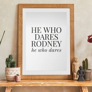 May include: A framed print with the text "HE WHO DARES RODNEY he who dares" in black lettering on a white background. The frame is a light brown wood and the print is sitting on a wooden table with a few plants and figurines.