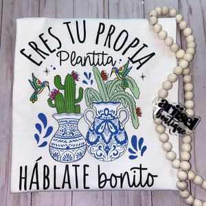 May include: White t-shirt with a black and white graphic design featuring two potted plants with hummingbirds and the text "Eres tu propia plantita. Hablate bonito".