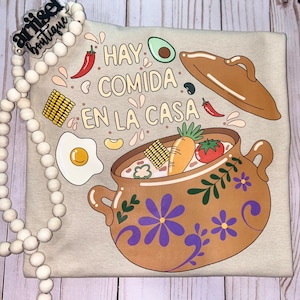 May include: A beige t-shirt with a brown pot of food and the text "Hay comida en la casa" in Spanish. The pot is decorated with purple flowers and green leaves. There are various food items inside the pot, including corn, carrots, tomatoes, and an egg.