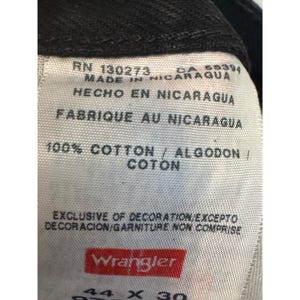 Op de afbeelding: Close-up van een wit label van een zwarte Wrangler-jeans. Het label bevat tekst in het Engels, Spaans en Frans, waaronder "100% Katoen" en "Gemaakt in Nicaragua". De maat is 44 x 30 inch.