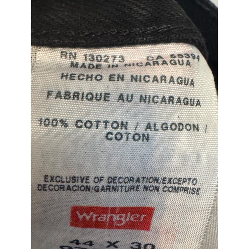 Op de afbeelding: Close-up van een wit label van een zwarte Wrangler-jeans. Het label bevat tekst in het Engels, Spaans en Frans, waaronder "100% Katoen" en "Gemaakt in Nicaragua". De maat is 44 x 30 inch.