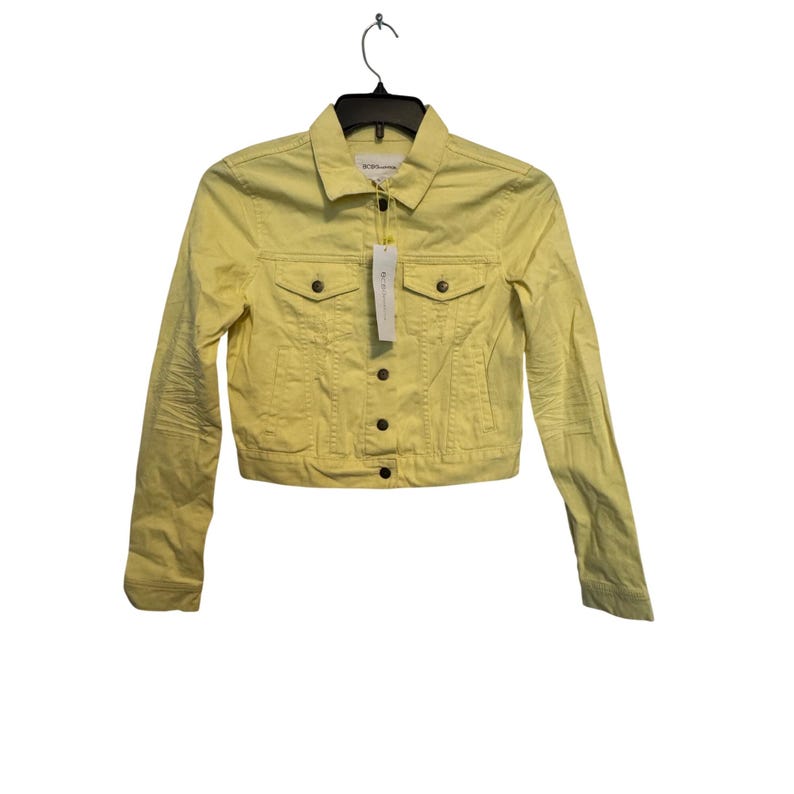 May include: A pale yellow denim jacket with a classic collar, two chest pockets, and button closures. The jacket is hanging on a black hanger and has a price tag attached. The jacket is a cropped style.