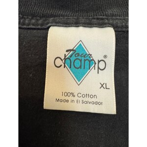 Puede incluir: Primer plano de una etiqueta de camiseta negra. La etiqueta presenta el texto "Tour Champ" en una fuente estilizada, con una forma de diamante turquesa. La etiqueta tambi&eacute;n dice "XL", "100% Algod&oacute;n" y "Made in El Salvador".